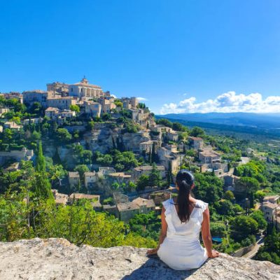 A young woman in a white dress sits peacefully on a rocky ledge, gazing at the picturesque village of Gordes. The bright sky enhances the lush greenery, Gordes Luberon Provence